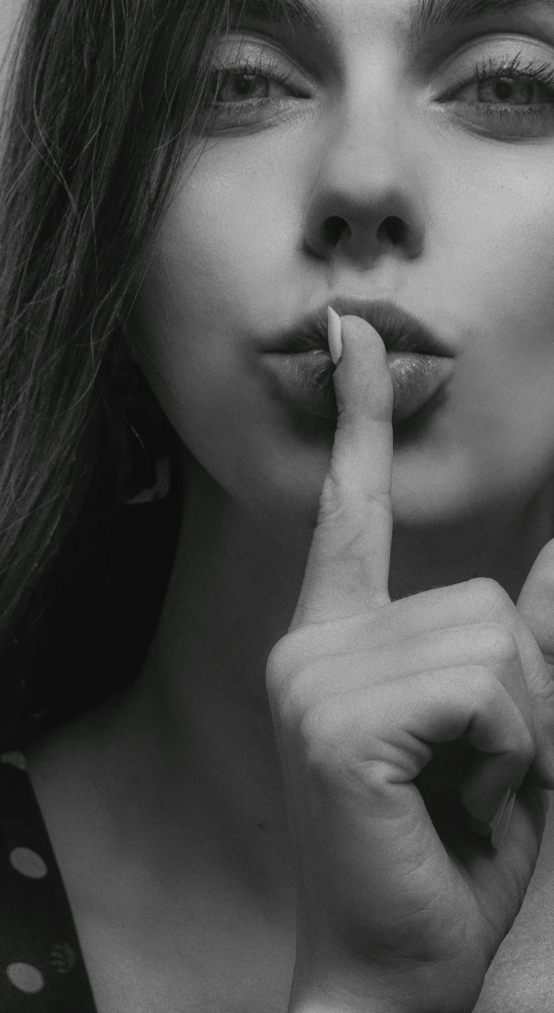 Close-up black and white portrait of a woman gesturing silence with a finger over lips.